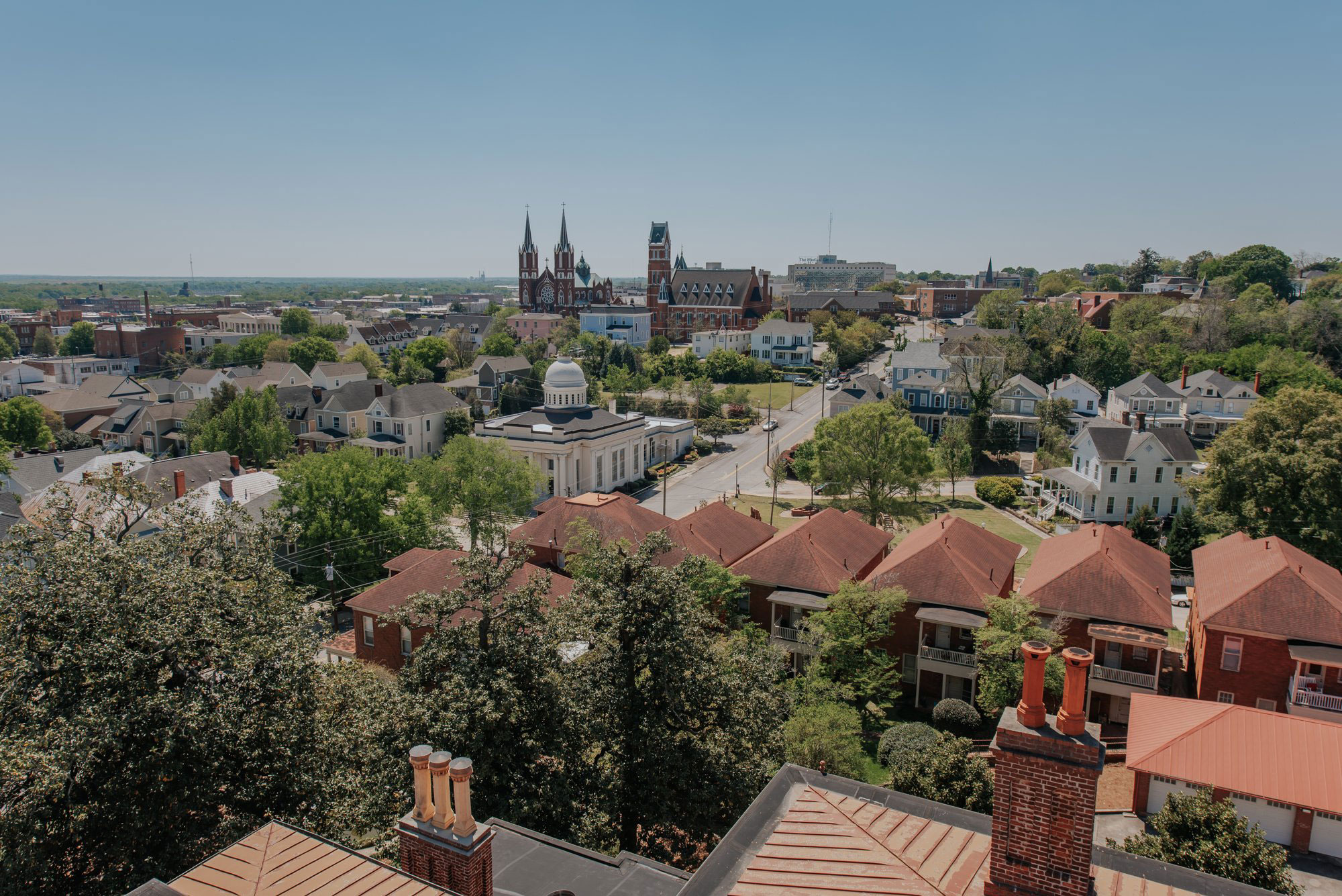 Aerial view of downtown Macon, Georgia. Near the bottom of the photo are a row of several red-brick houses intermingled with several green-leafed trees. There is a main street running across the photo and there are cars on the road. The sky above the whole scene, which includes lots of other old-fashioned buildings and dark-green trees, including a regal-looking cathedral, is dusty blue.