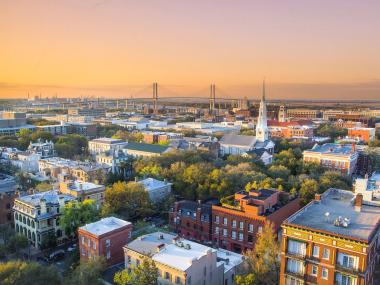 An aerial view of historic Savannah, Georgia. In the foreground are several antique brick buildings surrounded by trees among the streets; the lower half of the photo includes scenery like this, and along the horizon is the Talmadge Memorial Bridge. The sky is painted orange and purple by a Georgia sunset.
