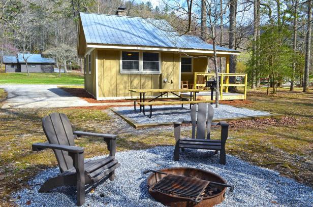 A cottage at Vogel State Park in Blairsville, Georgia, sits small and quaint in the middle of a grassy lawn with sparse trees littering the background. The cottage is painted a light yellow. In front of the cottage is a picnic table, and in front of that, at the very foreground, are two lawn chairs in front of a fire pit. The sky above the scene is blue and the sun shines through the branches of the surrounding trees.