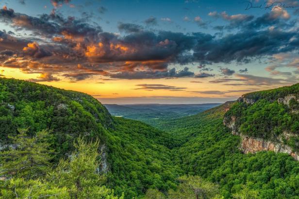 A spring sunset at Cloudland Canyon State Park in Rising Fawn, Georgia, glows orange and pink on the distant horizon. The sky above is dark, dusty blue and littered with dark-gray clouds. In the foreground is a valley covered in green trees.